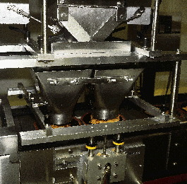 Near the head of the line, the black bowls in their carrier plates pass beneath the twin hoppers (left) of the filling station.
