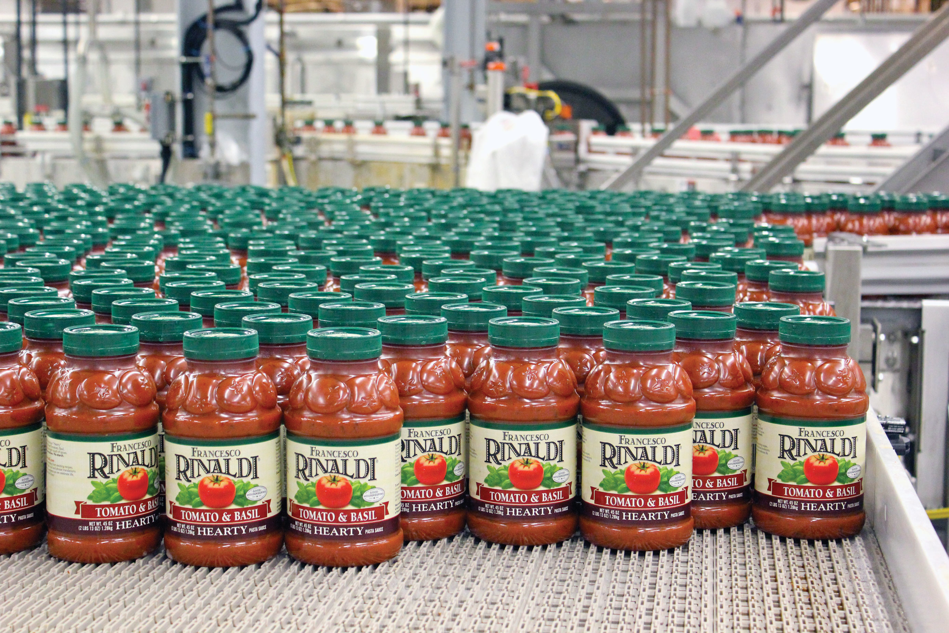 Pasta sauce bottles enter a labeler at left, then exit labeled at right. Filled, capped, and labeled bottles accumulate on a high-speed bottling line at LiDestri&rsquo;s Rochester, NY, facility.