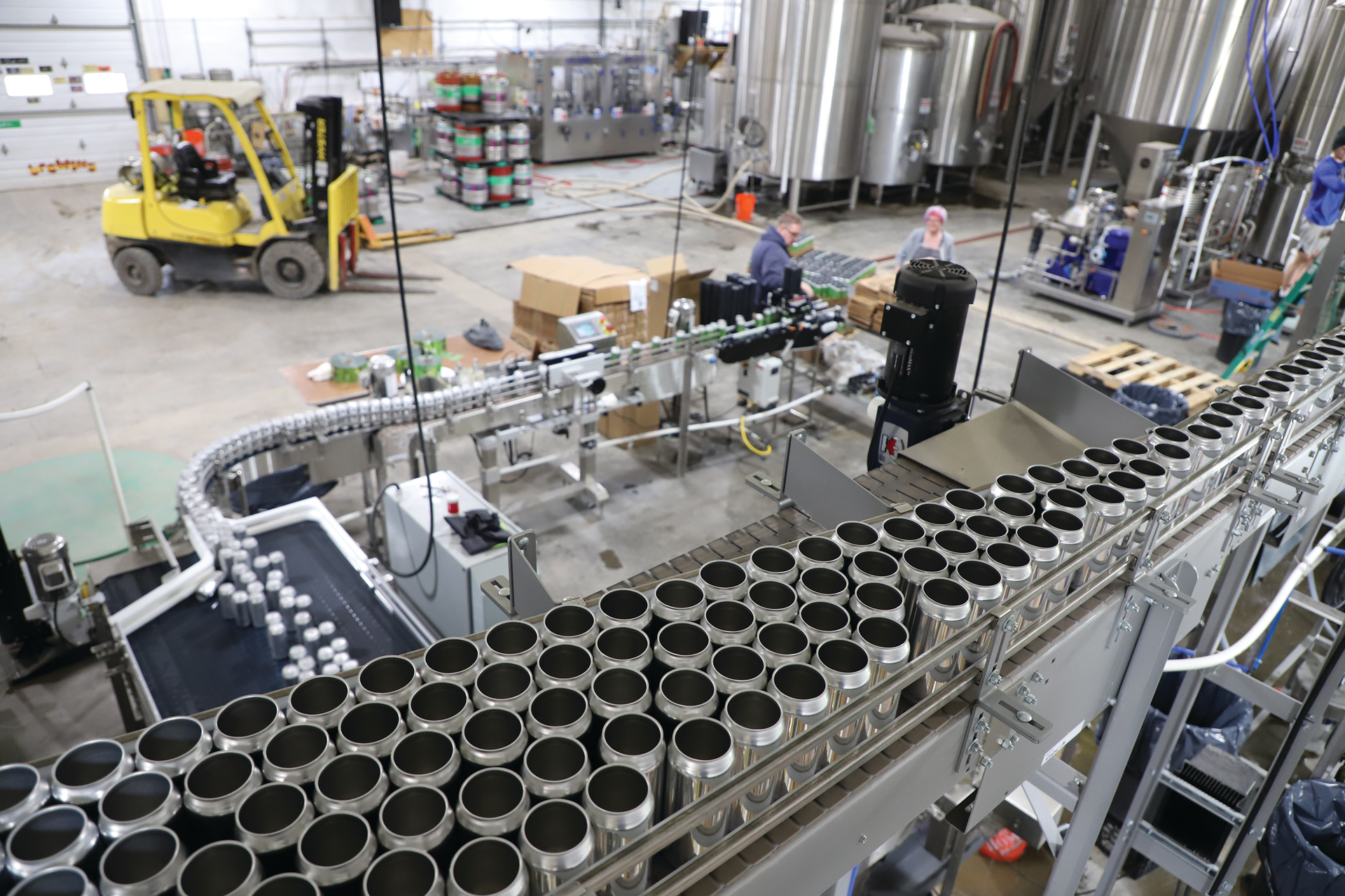 Brightstock cans are depalletized overhead on a high-trim Ska Can-i-Bus, then single-filed and lowered through a twist rinser on the way to the filler and seamer.