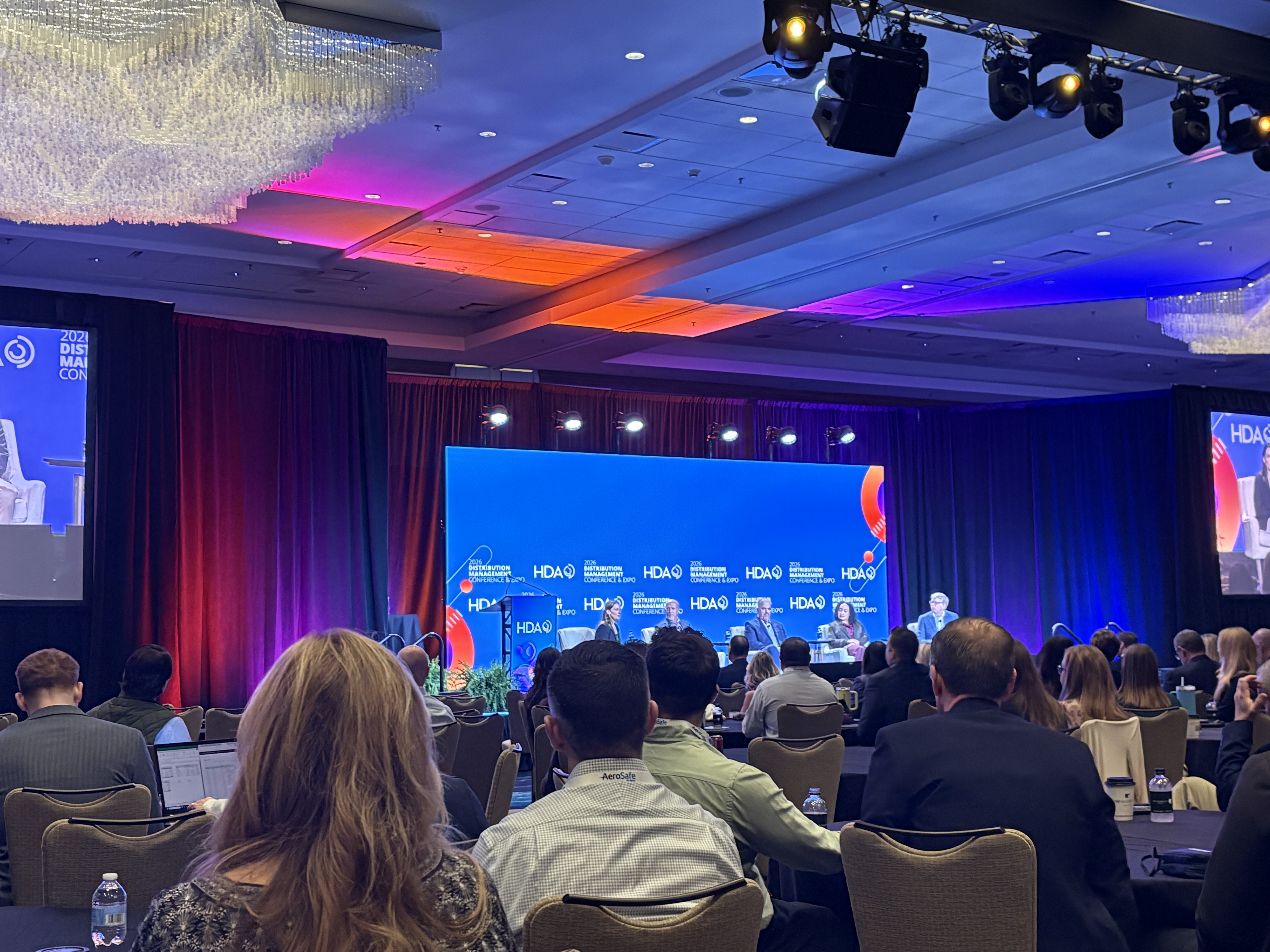 Audience members seated at a corporate conference event with blue and orange stage lighting and branded backdrop.