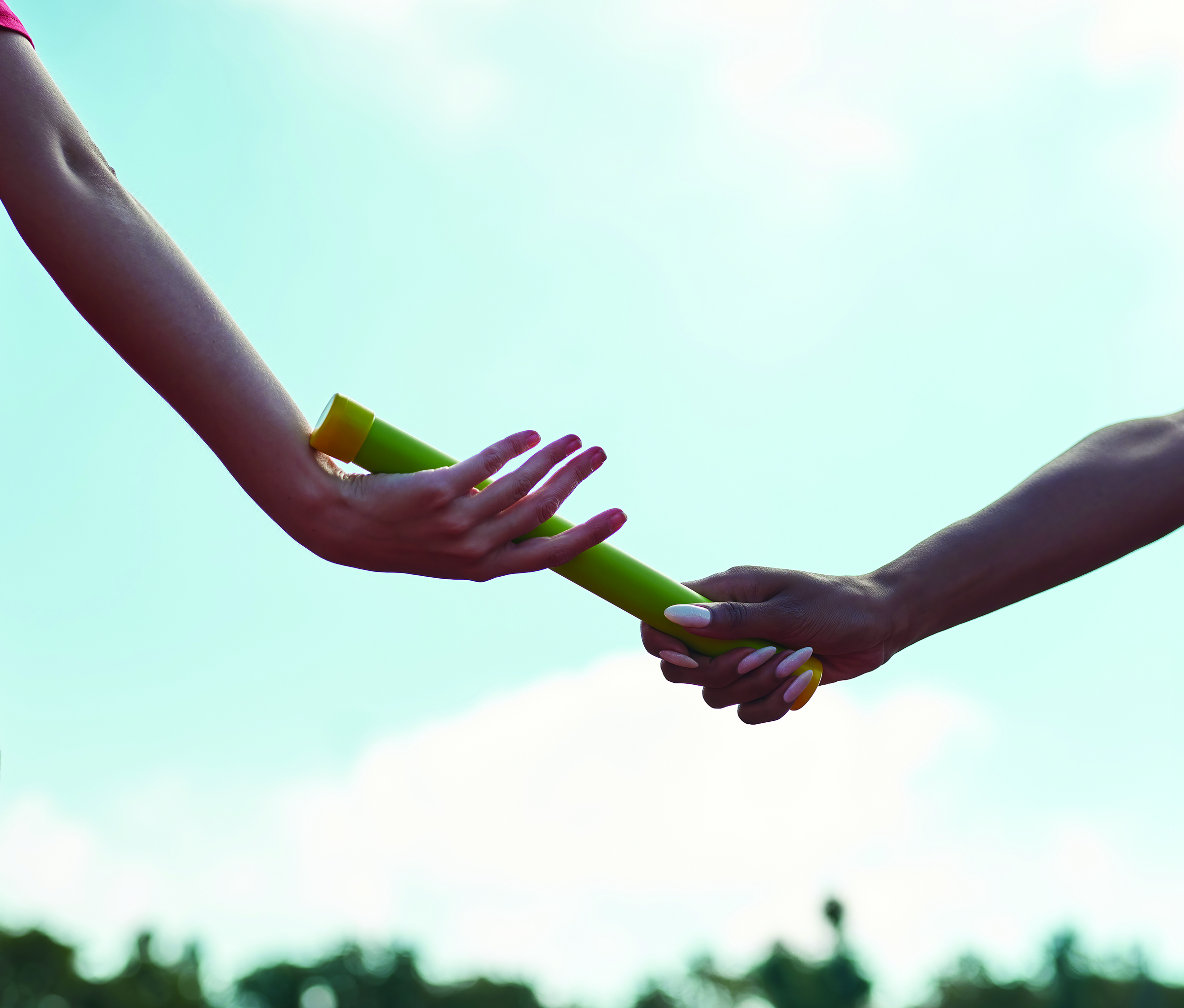 Two hands exchanging a green relay baton against a clear blue sky