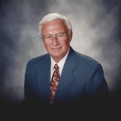Professional portrait of an elderly man in navy blazer and patterned tie against neutral background