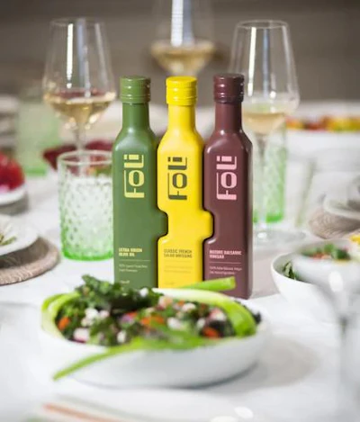 Three olive oil bottles with green, yellow, and burgundy labels displayed on elegant dining table with fresh salad and wine glasses