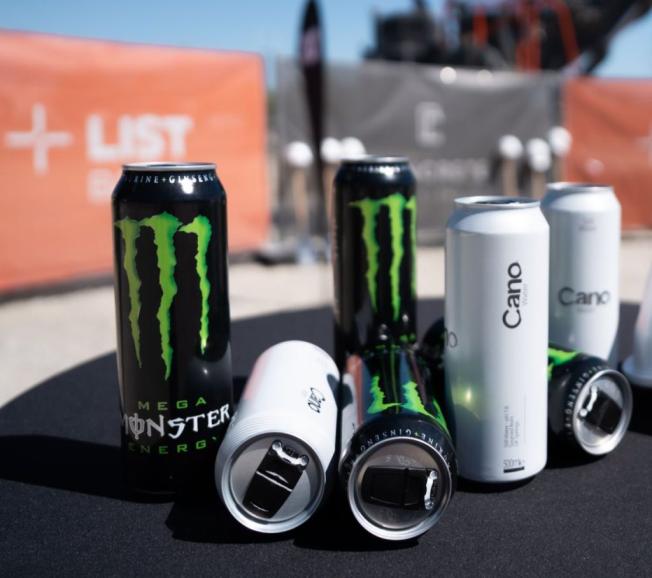Collection of Monster Energy drink cans and white beverage cans displayed outdoors with orange branded backdrop