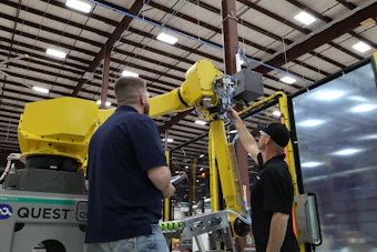 Two technicians service a yellow industrial robotic arm in a warehouse facility with metal framework ceiling.