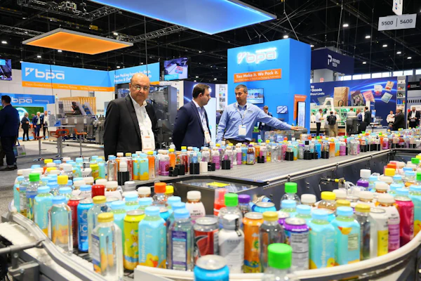 Three professionals examine colorful consumer product bottles displayed on curved tables at a trade show booth with blue and orange Bpda branding.