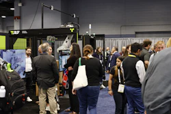 Attendees gather around exhibition booths at a trade show, with TRF branding visible on a black display booth featuring neon yellow accents.