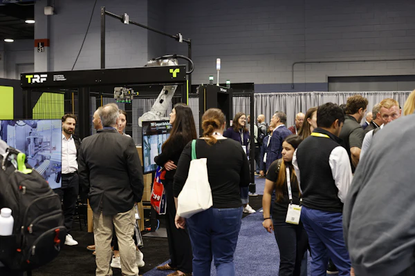 Attendees gather around exhibition booths at a trade show, with TRF branding visible on a black display booth featuring neon yellow accents.