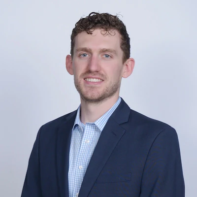 Professional headshot of a smiling man in navy blazer and light blue checkered shirt against white background