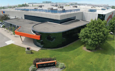 Aerial view of modern industrial facility with curved green building, white roofing structures, and orange entrance signage on manicured grounds