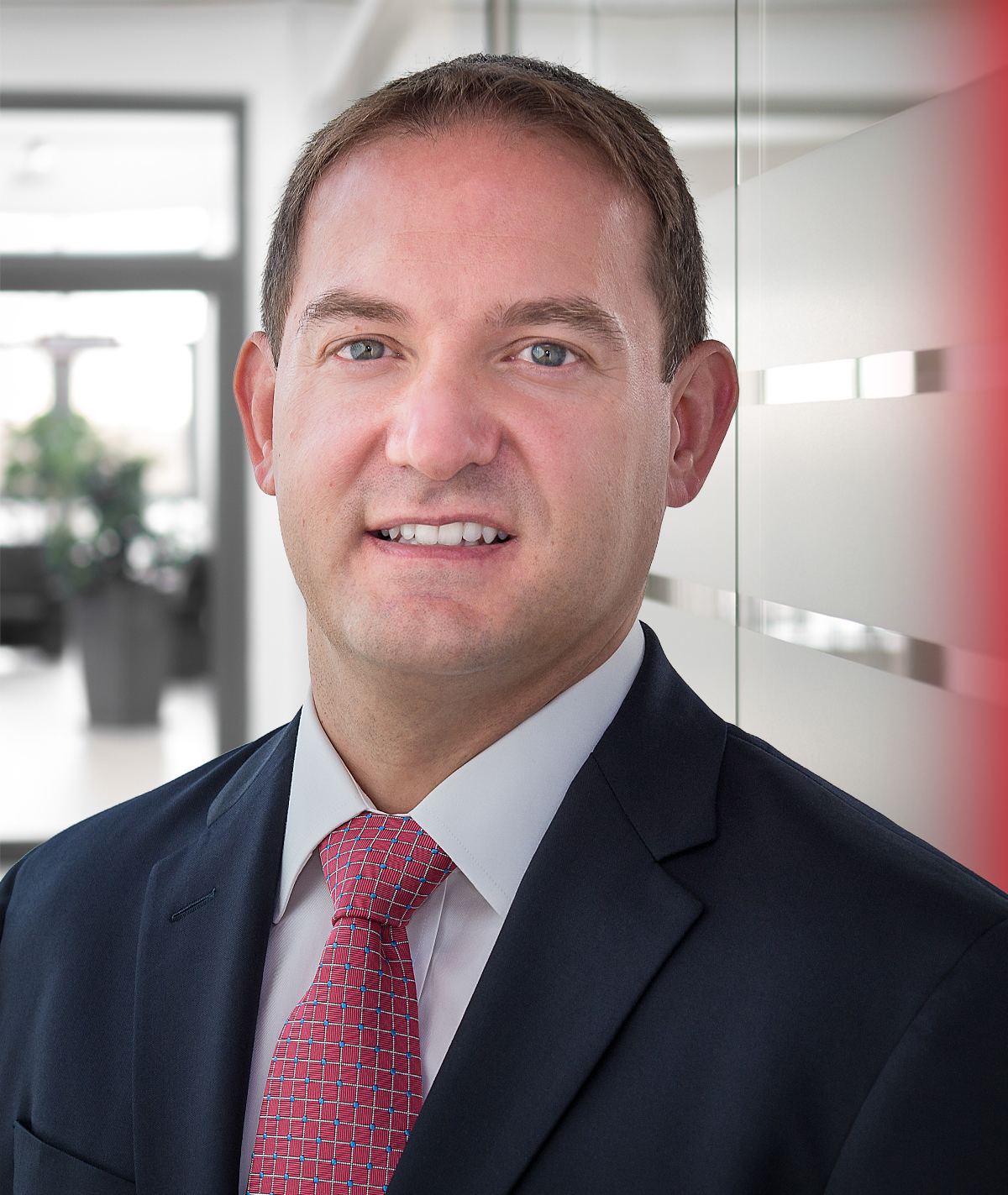 Professional headshot of a smiling man in navy blazer and red patterned tie in modern office setting